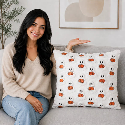 A smiling woman sits on a light gray sofa next to a white textured throw pillow featuring a repeating pattern of orange pumpkins and white ghosts with black eyes.