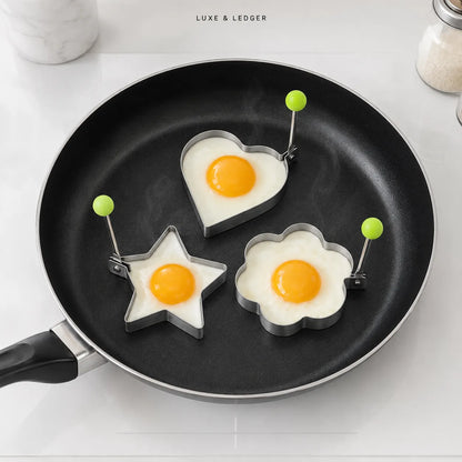 Three metal egg-shaped frying mold pans shaped like a heart, star, and flower, each holding a fried egg, set inside a black non-stick frying pan on a white countertop. The text 'LUXE & LEDGER' is visible at the top.
