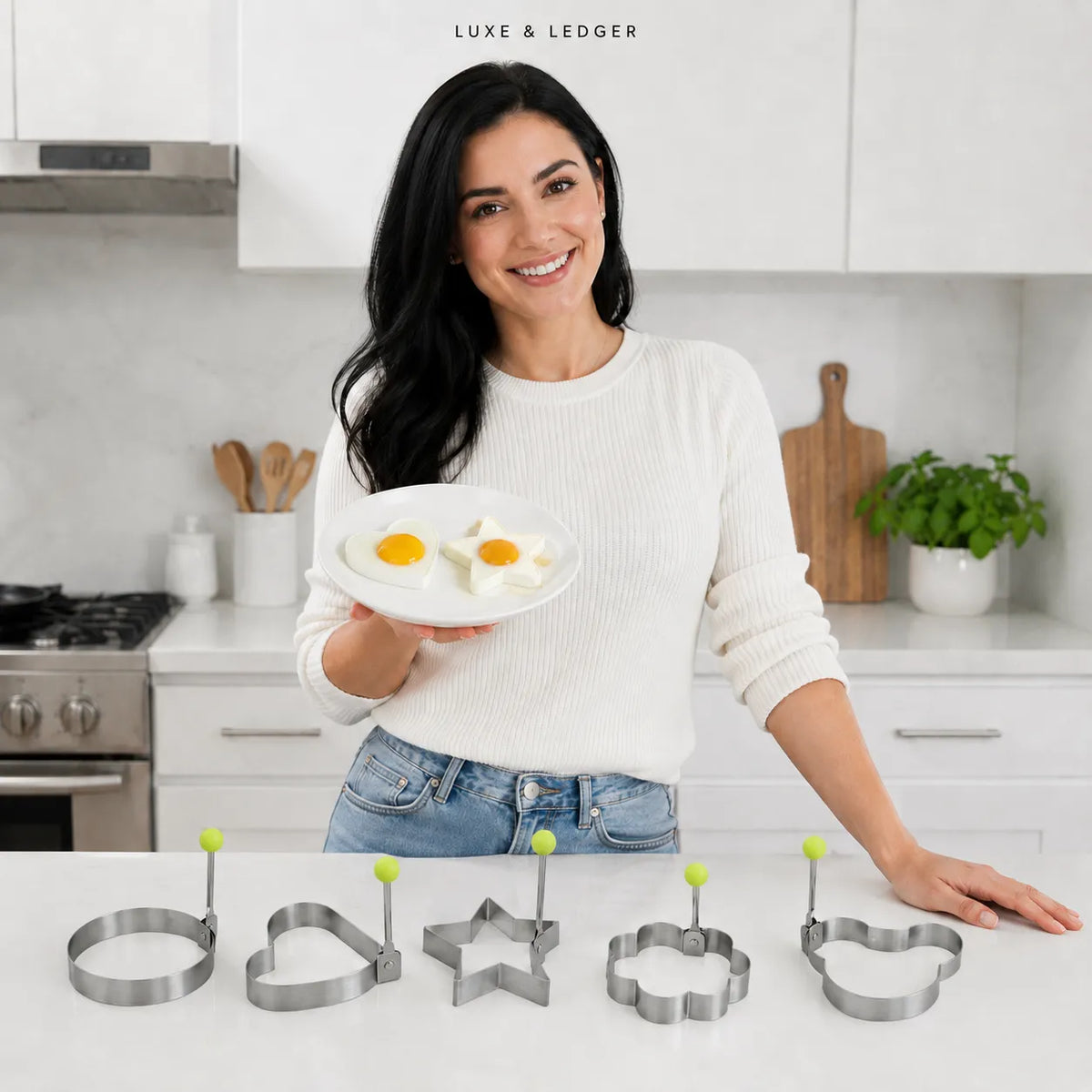 A woman in a white sweater and jeans stands in a bright kitchen holding a plate with two eggs, with five metal egg-casserole mold cutters lined up on the counter in front of her. The cutters are shaped like a ring, heart, star, flower, and fish.