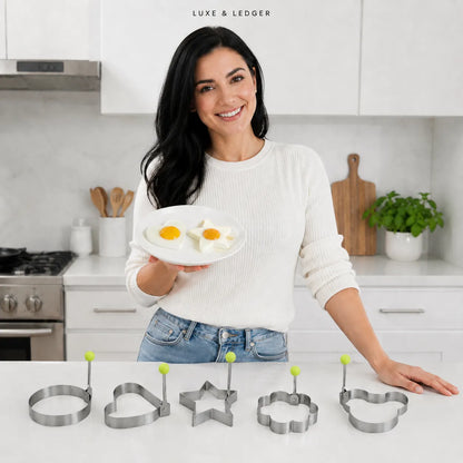 A woman in a white sweater and jeans stands in a bright kitchen holding a plate with two eggs, with five metal egg-casserole mold cutters lined up on the counter in front of her. The cutters are shaped like a ring, heart, star, flower, and fish.