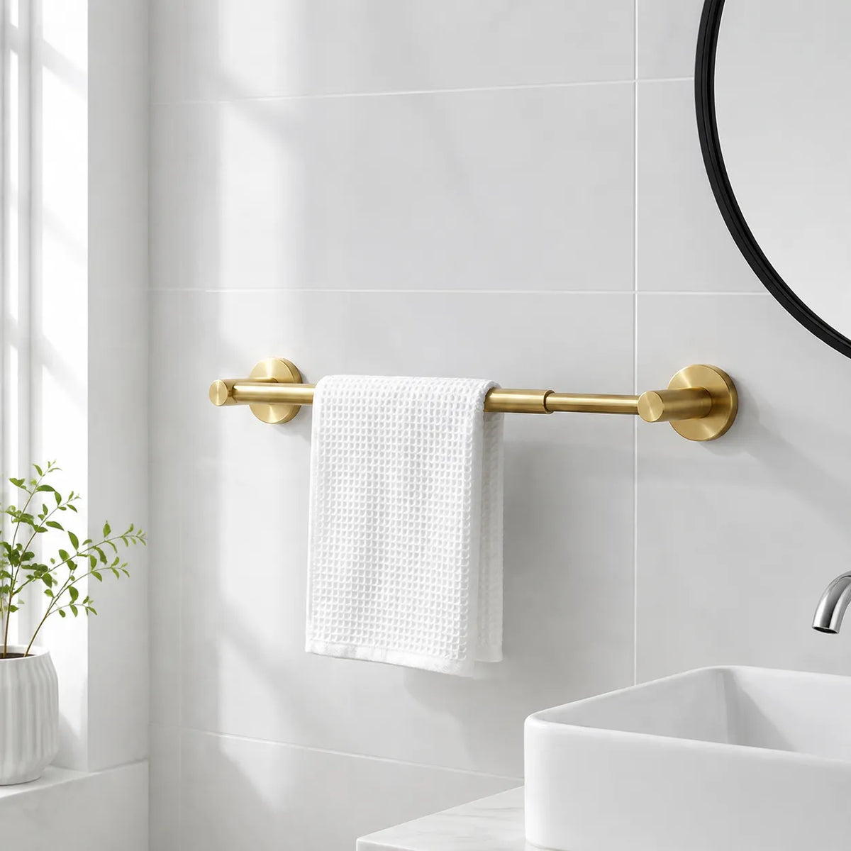 A matte gold double towel bar mounted on a white tiled bathroom wall, holding a white textured towel. A white vessel sink, a small potted plant, and part of a round mirror are visible in the scene.