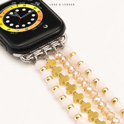 A close-up of a black smartwatch with a decorative multi-strand beaded strap. The strap features pale pink round beads, faceted gold beads, and gold clover-shaped charms.