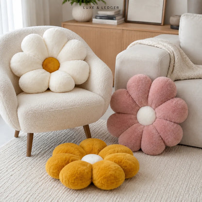 Three plush flower-shaped throw pillows in cream, dusty pink, and mustard yellow are arranged in a cozy living room setting next to a beige armchair and sofa.