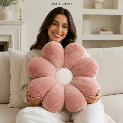 A smiling woman sits on a cream sofa holding a large plush flower-shaped pillow in dusty pink with a white center. The background shows a bright, modern living room with shelves and a fireplace.