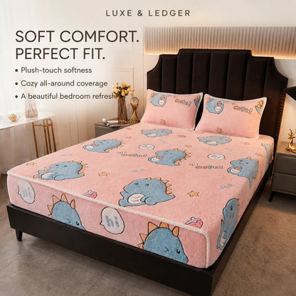 A pink bedding set with a cartoon blue dinosaur pattern is shown on a bed in a styled bedroom. The set includes a fitted sheet and two matching pillowcases.