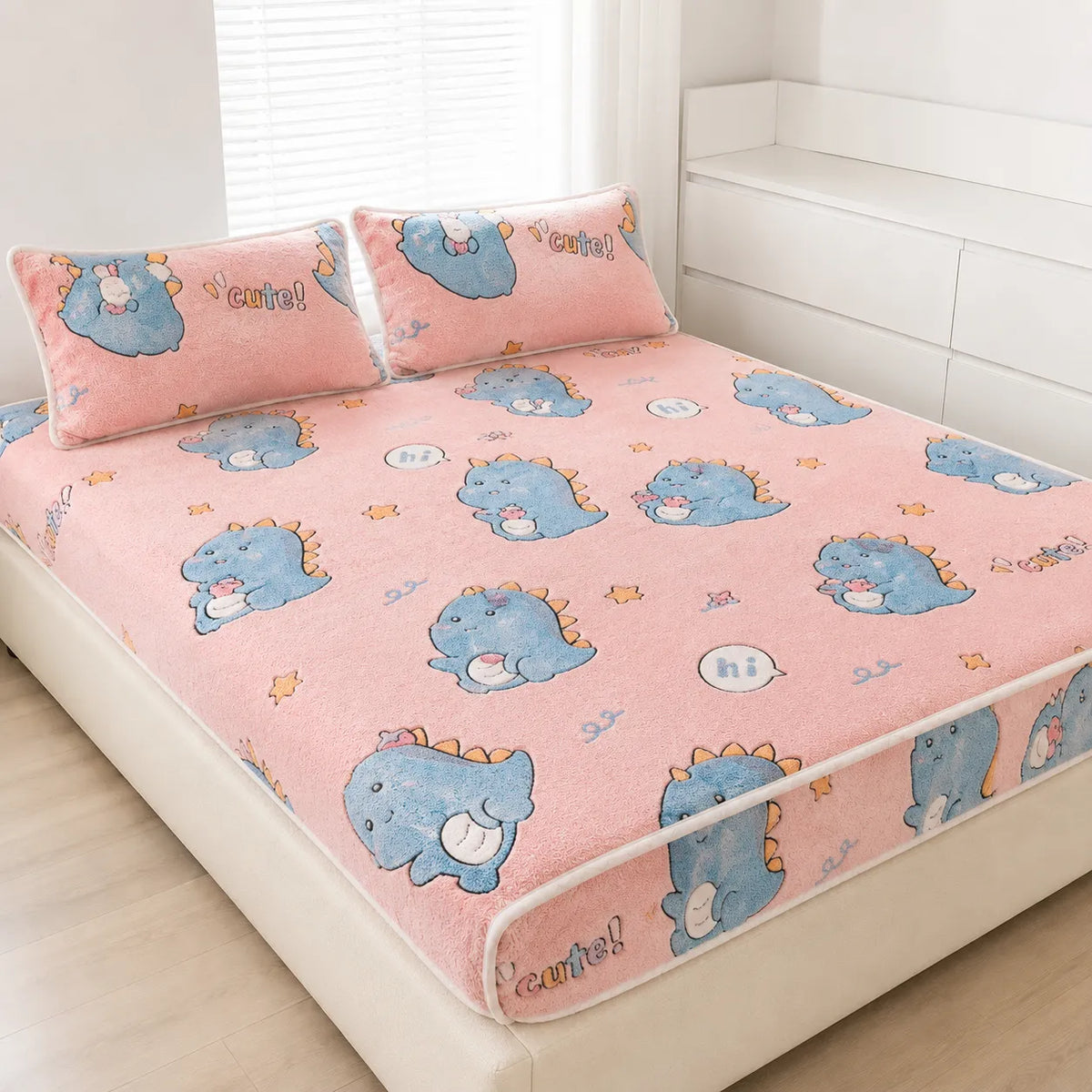 A pink bedspread and matching pillowcases featuring a cute blue dinosaur cartoon print, styled on a bed in a bright, modern bedroom.