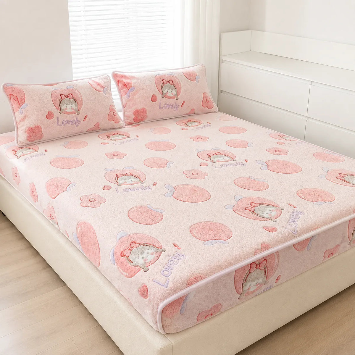 A pink bed sheet set with two matching pillowcases on a bed. The sheet and pillows feature a cute cartoon cat character inside a pink heart, surrounded by pink peach-like circles and small flowers, with the word 'Lovely' printed in blue.