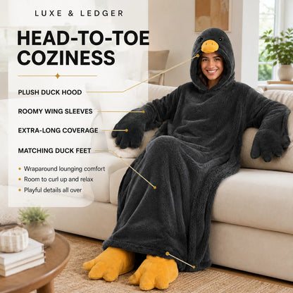 A person reclining on a beige sofa wearing a plush duck-themed head-to-toe cozy blanket with a duck hood, wing sleeves, matching foot covers, and yellow-orange beak and feet details. The image includes marketing text and callout lines explaining the product features.