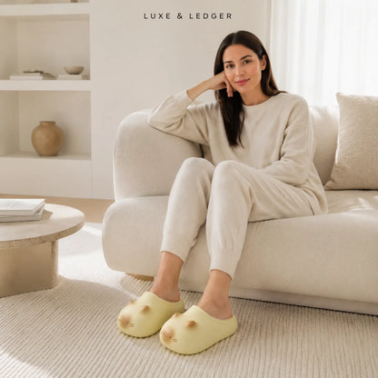 A woman sits on a cream-colored sofa in a bright, modern living room, wearing a matching beige lounge set and pale yellow plush slip-on slippers with cat ears. The brand name 'LUXE & LEDGER' is displayed at the top of the image.