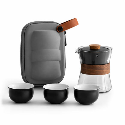 A tea brewing set on a white background, including a gray hard case with a brown leather strap, a glass teapot with a black lid and brown wood accent, and three black tea cups with white interiors.