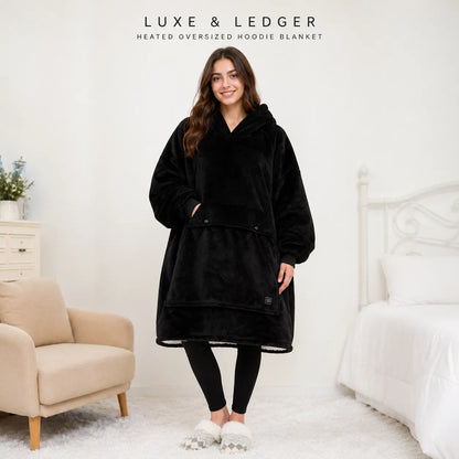 A woman stands in a bedroom wearing a black Luxe & Ledger heated oversized hoodie blanket. The image includes the brand text 'LUXE & LEDGER HEATED OVERSIZED HOODIE BLANKET' at the top.