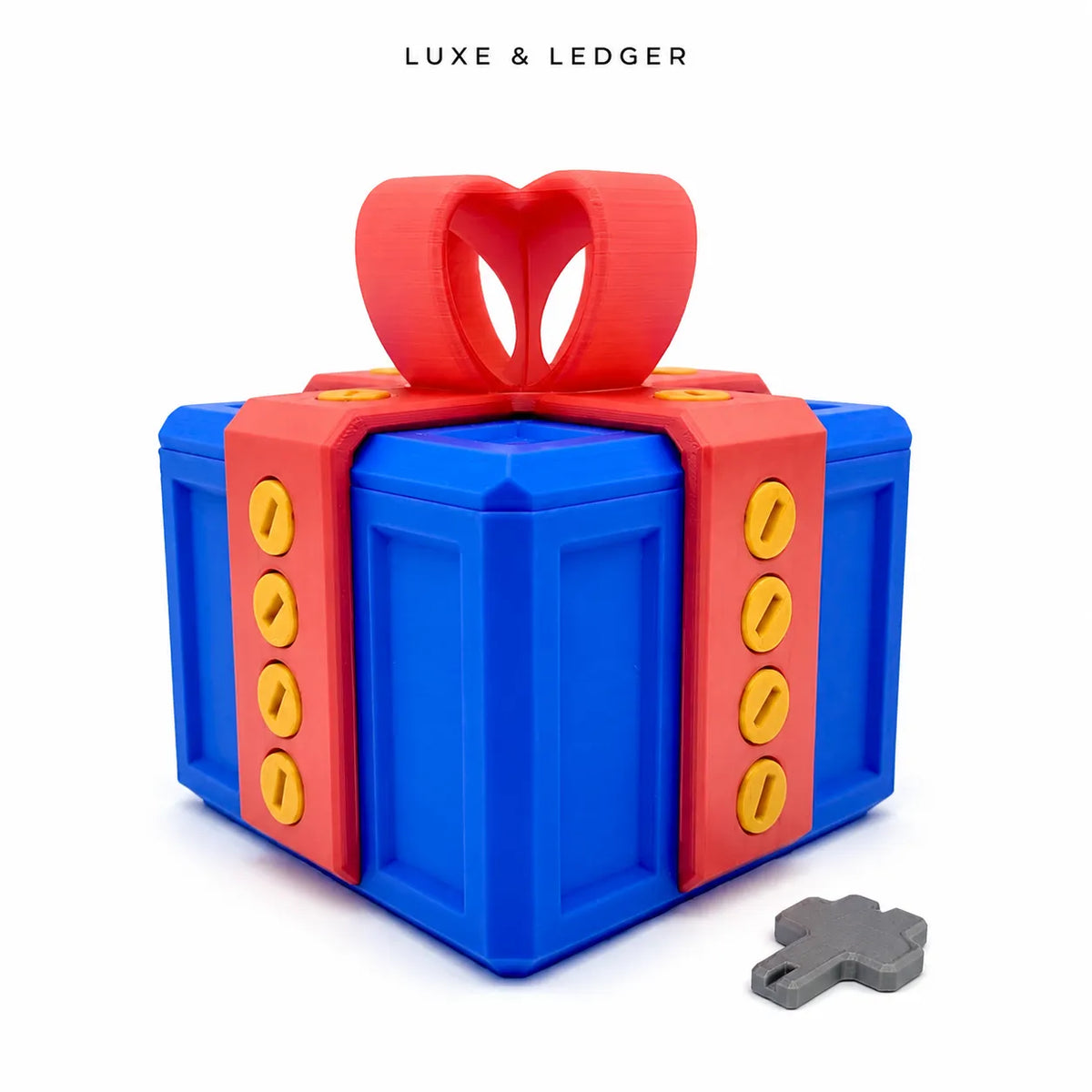 A 3D-printed gift box toy in blue and red with yellow button details, featuring a red heart-shaped bow on top and a small gray key in the foreground. The brand name 'LUXE & LEDGER' is printed at the top of the image.