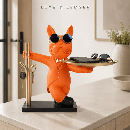 An orange cat figurine wearing sunglasses and a bow tie, holding a tray with a wallet and sunglasses, and standing next to a gold key rack on a black base. The figurine is placed on a light beige countertop in a modern interior setting.