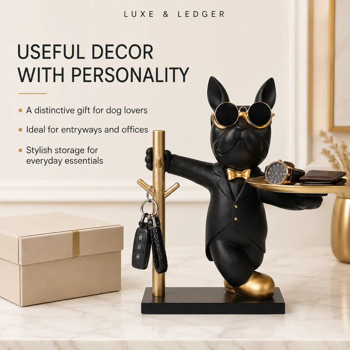 A black French bulldog figurine wearing gold sunglasses, a gold bow tie, and a black tuxedo, standing on a black base and holding a gold tray with a watch and wallet. The figurine is next to a gold key holder with keys, set on a marble surface with a beige gift box.