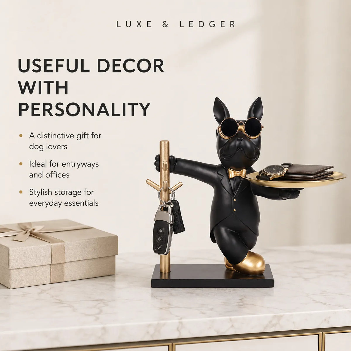 A black French bulldog key holder figurine wearing gold sunglasses, a bow tie, and gold shoes, standing on a black base and holding a tray with accessories while holding a key on a gold key rack. The figurine is displayed on a marble tabletop next to a wrapped gift box, with promotional text on the left side.