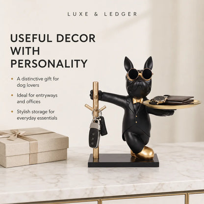 A black French bulldog key holder figurine wearing gold sunglasses, a bow tie, and gold shoes, standing on a black base and holding a tray with accessories while holding a key on a gold key rack. The figurine is displayed on a marble tabletop next to a wrapped gift box, with promotional text on the left side.