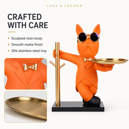 An orange cat figurine wearing a gold suit, sunglasses, and bow tie, holding a round gold tray while standing on a black base next to a gold coat rack. The image includes marketing text, two inset detail photos, and the Luxe & Ledger brand name at the top.