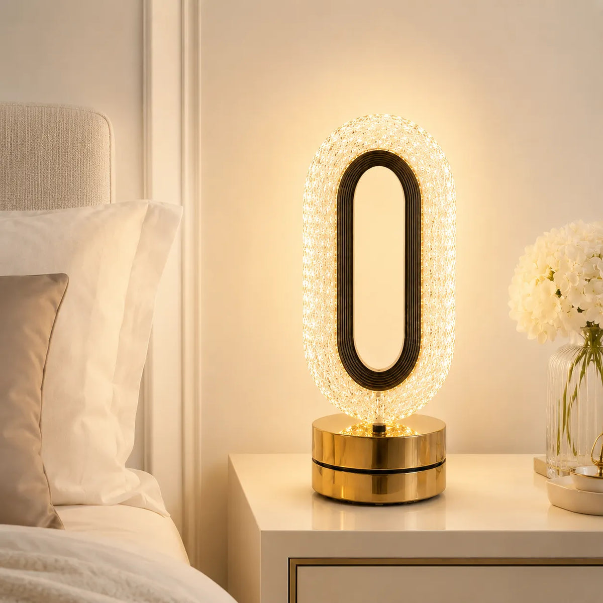 A modern oval table lamp with a black frame and a gold base, encased in a clear crystal-like border that glows warmly, placed on a white bedside table in a bedroom setting.