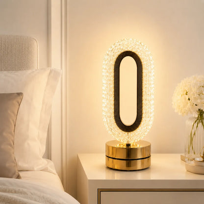 A modern oval table lamp with a black frame and a gold base, encased in a clear crystal-like border that glows warmly, placed on a white bedside table in a bedroom setting.