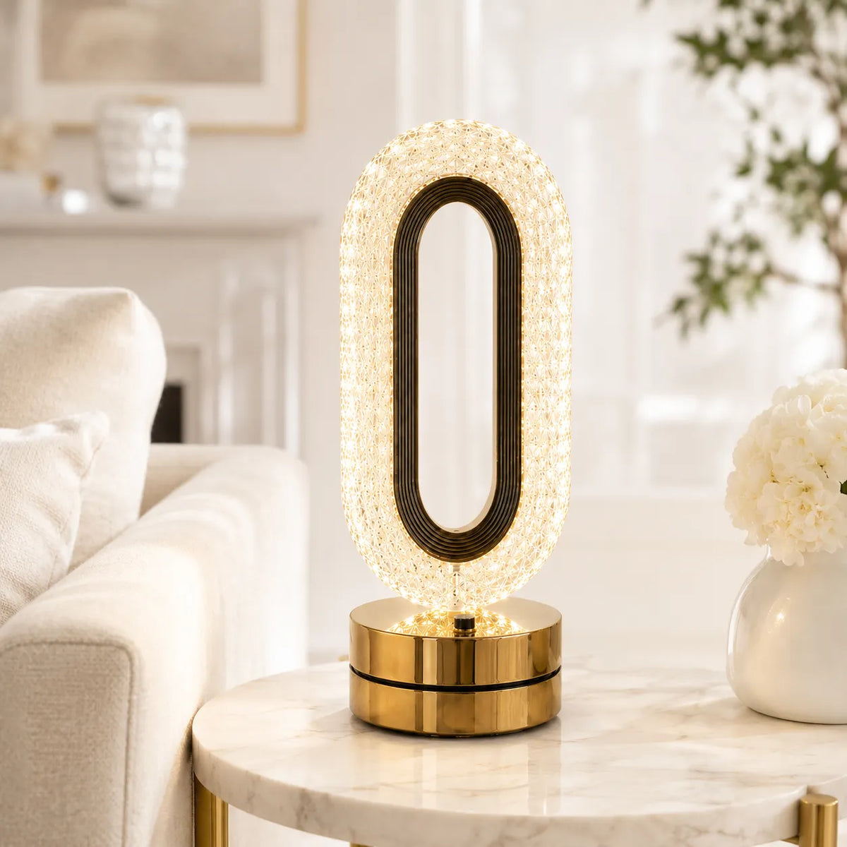 A modern oval table lamp with a gold base and a clear faceted crystal-like outer frame, shown lit on a marble side table in a bright living room setting.