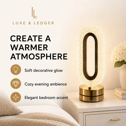 A Luxe & Ledger table lamp with an oval crystal-encrusted frame and a polished gold base, styled on a white bedside table next to a bed. The image includes promotional text highlighting its warm glow and cozy ambiance.