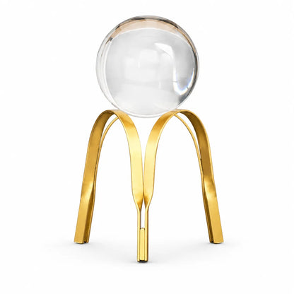A modern decorative sculpture featuring a clear glass sphere balanced on a polished gold metal tripod base, set against a solid white background.