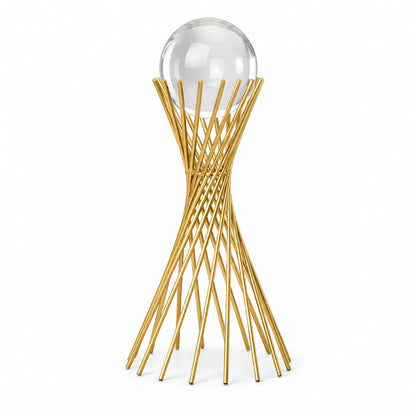 A modern gold metal sculpture stand with a twisted, lattice-like base and a clear glass sphere resting on top, set against a plain white background.