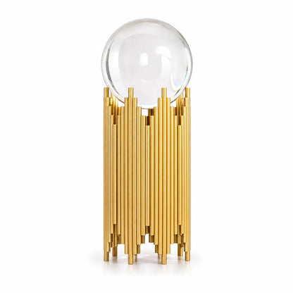 A modern table lamp featuring a clear glass crystal sphere on a metallic gold stand composed of vertical cylindrical rods, set against a plain white background.