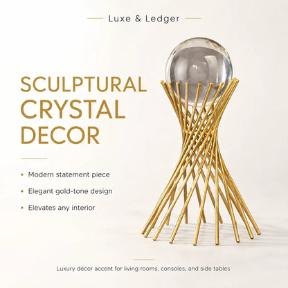 A luxury crystal decor sculpture featuring a clear glass sphere resting on a gold-tone twisted metal stand, presented on a clean off-white background with promotional text reading 'Luxe & Ledger' and 'Sculptural Crystal Decor'.