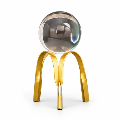 A modern decorative sculpture featuring a clear glass sphere balanced on a polished gold metal stand with three curved legs, set against a plain white background.