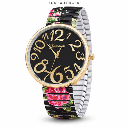A wristwatch with a black dial, gold-tone numerals and hands, a gold-tone bezel, and a silver-tone metal link bracelet. The bracelet has a black background with a pink and green floral print.