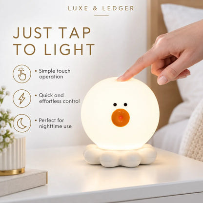 A cute duck-shaped night light by Luxe & Ledger, glowing with a warm white light on a bedside table. A hand is touching the top of the glowing sphere to demonstrate the touch-sensitive operation.