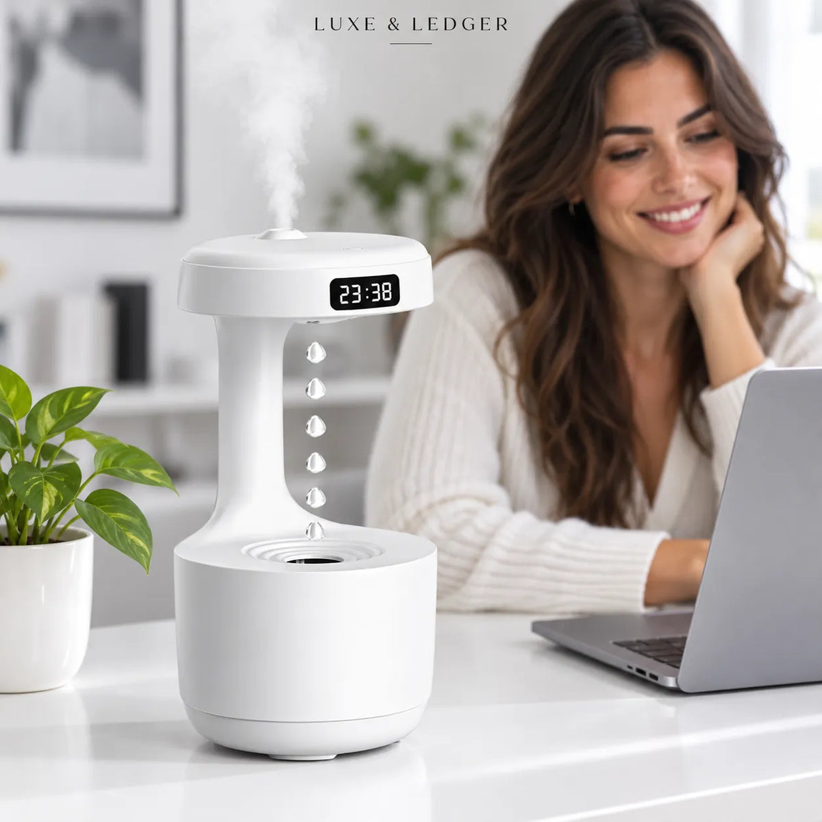 A white tabletop humidifier with a digital display reading 23:38 and misting at the top, shown on a desk in a bright home office setting with a woman working on a laptop in the background. The brand name 'LUNE & LEDGER' is visible at the top.