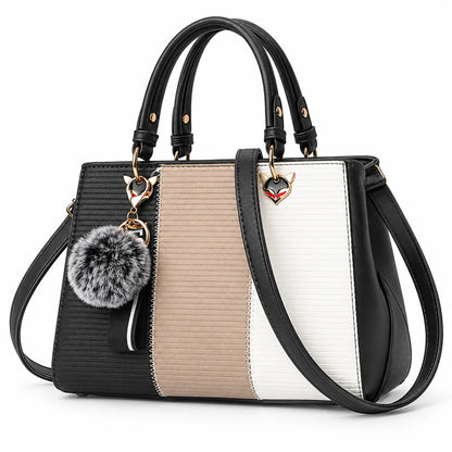 A structured handbag with a color-blocked design in black, beige, and white textured panels, featuring black leather handles, a detachable shoulder strap, and a gray faux-fur pom-pom charm with a gold fox-head accent.