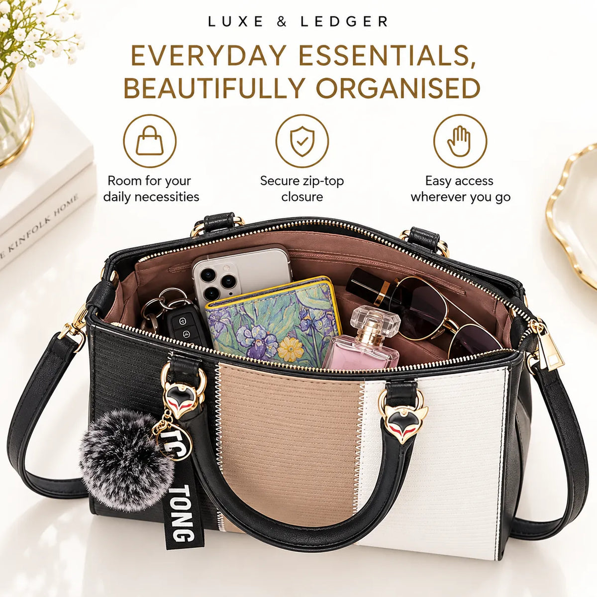 A structured tote handbag shown open, filled with everyday essentials like a phone, wallet, sunglasses, and perfume. The bag has a color-blocked design in black, beige, and white with gold-tone hardware and a gray pom-pom charm.