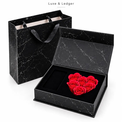 A luxury gift set featuring a heart-shaped arrangement of preserved red roses inside an open black marble-patterned box, with a matching black marble gift bag and a branded 'Luxe & Ledger' box behind it, on a white background.