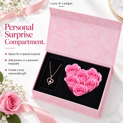 A pink presentation box with a black interior, shown open on a light marble surface. Inside are a pink heart-shaped rose arrangement and a gold heart pendant necklace.