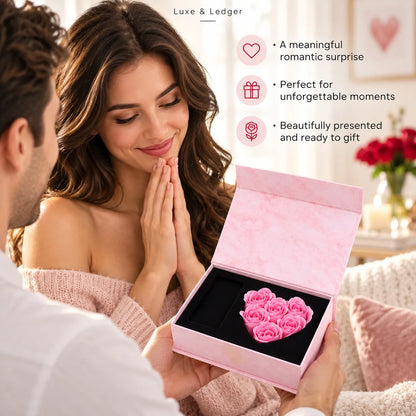 A man presents a pink heart-shaped rose gift box to a woman in a cozy bedroom setting. The box is open, revealing a cluster of small pink roses on a black insert.