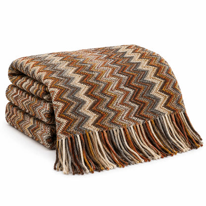 A folded woven throw blanket with a zigzag chevron pattern in earthy tones of brown, beige, rust orange, and gray, finished with a fringed edge, shown on a plain white background.