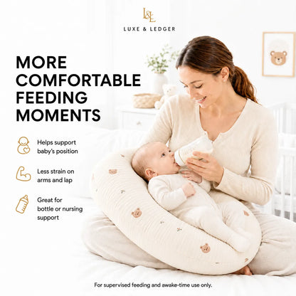 A woman sits on a bed holding a baby on a cream-colored nursing pillow while feeding the baby with a bottle. The image includes text highlighting the benefits of the nursing pillow from Luxe & Ledger.