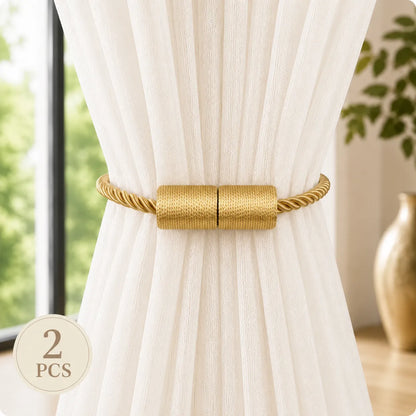 A pair of gold curtain tiebacks with twisted rope sides and a textured cylindrical center, shown tied around a white curtain in a bright room. A circular '2 PCS' badge is in the lower-left corner.