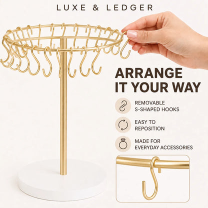 Gold jewelry stand with a white circular base and multiple S-shaped hooks, shown with a hand adjusting a hook. Text reads 'LUXE & LEDGER' and 'ARRANGE IT YOUR WAY' with feature icons.