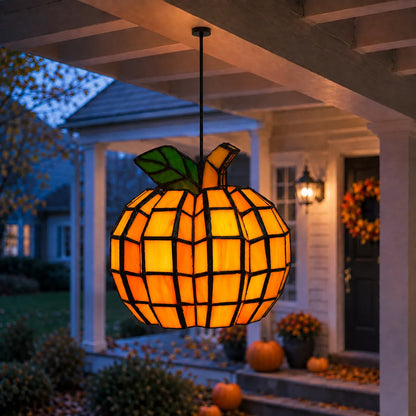 A hanging pumpkin-shaped pendant light with a stained-glass style orange and yellow body, black outlines, and green leaves, suspended over a porch at dusk.