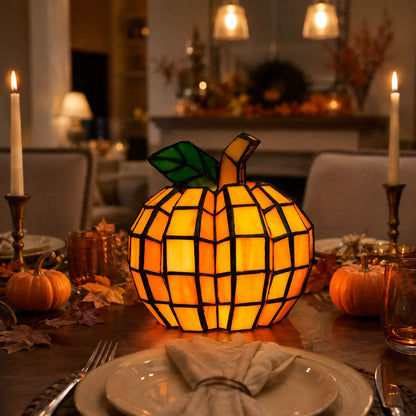 A stained-glass pumpkin table lamp glowing with warm orange and yellow light, set on a decorated dining table with candles, pumpkins, and autumn leaves in a cozy indoor setting.