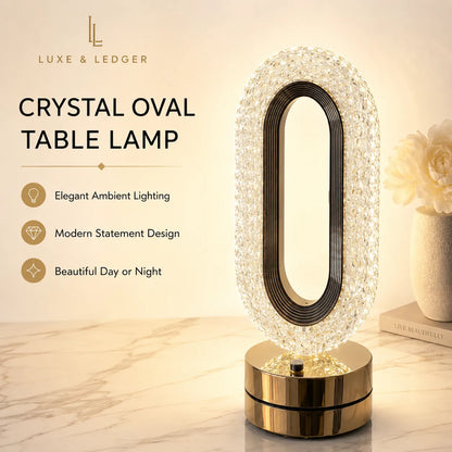 Luxe & Ledger crystal oval table lamp with a faceted clear crystal outer frame, black inner ring, and a shiny gold cylindrical base, shown on a marble surface with warm ambient lighting.