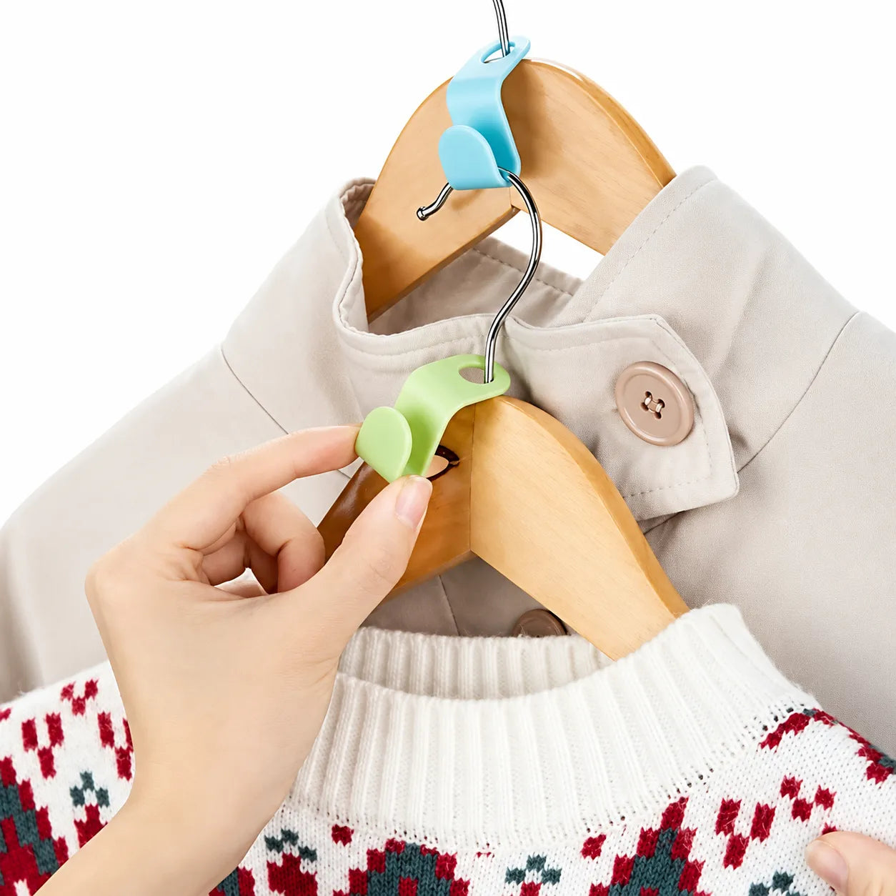 A hand is shown using a light green plastic clothes hanger hook clip to hang a white knit sweater with a red and green patterned design on a wooden hanger, next to a beige jacket on another hanger.