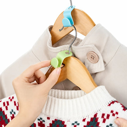 A hand is shown using a light green plastic clothes hanger hook clip to hang a white knit sweater with a red and green patterned design on a wooden hanger, next to a beige jacket on another hanger.