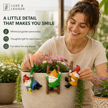 A smiling woman in a beige T-shirt holds a beige planter pot decorated with three whimsical garden gnome figures climbing or hanging on the front. The planter is filled with pink and white flowers, and green foliage drapes down.