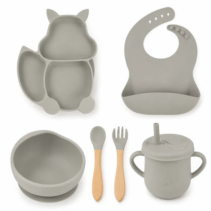 A baby feeding set in a uniform light gray silicone material, featuring a cat-shaped divided plate, a matching silicone bib, a bowl with a suction base, a lidded sippy cup with a straw, and a wooden spoon and fork.