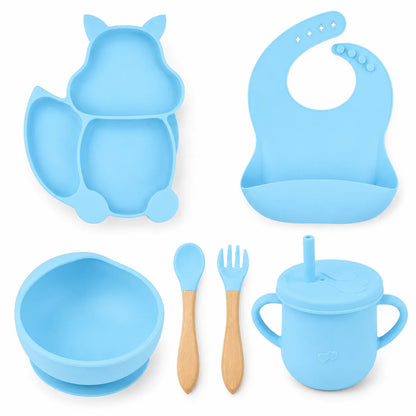 A light blue silicone baby feeding set on a white background, including a cat-shaped divided plate, a bib, a bowl, a sippy cup with lid and straw, a spoon, and a fork with wooden handles.